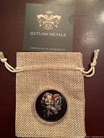 Photo showing the silver coin inside a small beige drawstring pouch with visible texture and a certificate of authenticity from Outlaw Metals behind it.