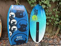 Two skim boards side by side against a wooden fence with some greenery visible.