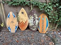 Four wooden skim boards standing upright on gravel against a fence with ivy, showing various designs and wear.