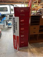 Front view of red filing cabinet with Coca-Cola logo decals on six drawers and brass handles, standing beside the Coca-Cola metal sign trimmed in white and red with 'refreshment centre' text.