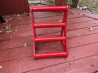 Red metal heavy duty wheel chock with triangular side supports and three horizontal cylindrical bars, placed on a wooden deck outside.
