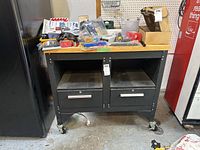 Front view of black metal frame work bench with natural wood top, two drawers, two open cubby spaces, and casters on wheels.