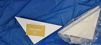 Two white triangular corner floating shelves, one wrapped in plastic, placed on a blue background.