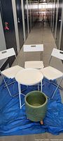 Three white Ikea folding chairs arranged around a small round white table with a green ceramic plant holder in front placed on a blue tarp.