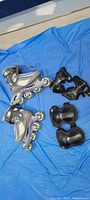 Photo showing pair of grey and silver inline roller blades with wheels and included black protective knee and elbow pads on blue background.