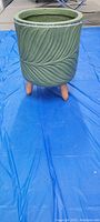 Front view of olive green ceramic plant stand with raised leaf design and wooden legs on blue tarp.