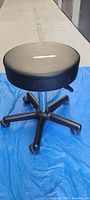 Black adjustable stool with five casters, round seat seen from side angle on blue tarp