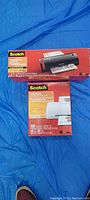 Photo of 3M Scotch thermal laminator in box and box of 100 thermal laminating pouches laid flat on blue background.