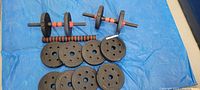 Two adjustable dumbbell bars with black and red foam grips, multiple cast iron weight plates arranged on a blue tarp.
