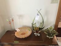 Photo showing two tall stemware glasses, one large ruffled glass decorative dish, and two art glass sculptures on a wood table.