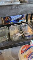 Photo showing multiple stacks of clear rectangular glass baking pans of various sizes, some nested inside each other, placed on a metal shelf.