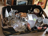 Box containing multiple candle holders of different sizes and materials including glass and metal, a small ceramic bowl, mug, and metal art pieces.