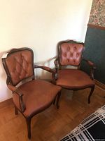 Two brown upholstered tufted parlour arm chairs with wood curved arms and legs, placed side by side on a wooden floor.