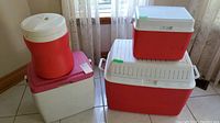 Four items arranged in a cluster comprising three coolers of varying sizes and a red thermos jug with a white lid, placed on tile flooring near a window with sheer curtains.