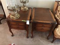 Pair of wooden end tables with graduated coloring, inlay look tops, two drawers each, and curved legs photographed side by side with decorative lamps on top.