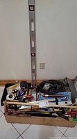Photo showing a large box level leaning against the wall with a box containing assorted hand tools including hammers, saws, and rollers.
