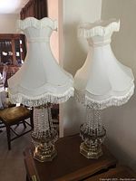 Pair of vintage lamps with clear glass bases and white fringed fabric shades on a wooden table near a wall.