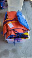 Four assorted life jackets stacked and placed in and on a plastic bin, showing colors and styles.