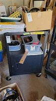Front view of black metal service cart with lower storage cabinet, caster wheels, and an electrical power strip attached on the top shelf side. Various miscellaneous items are on top, not included.