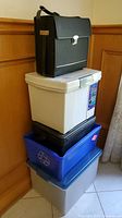 Stacked office-related storage items: black briefcase organizer, white plastic bin, black bin, blue recycling bin, and gray bin