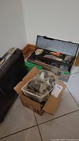 Photo showing metal tool box open with assorted tools and hardware, black plastic hard case adjacent, boxes and basket with casters in foreground.