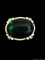 Close-up front view of the brooch showing large central faceted emerald green rhinestone crystal, surrounded by smaller round green rhinestones and clear rhinestones set into gold-tone metal.