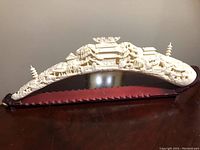 Side angle of carved faux ivory tusk on wooden stand showing detailed bridge and buildings.