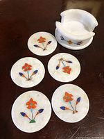 Top view of five round white marble coasters each with colorful pietra dura floral inlay arranged on a dark wooden table and coaster holder placed behind.