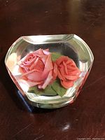 Clear faceted Lucite paperweight with two detailed pink roses and green leaves encased inside, resting on a wooden surface.