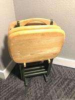 Four collapsible solid oak dinner tables resting on and stored in a green wooden stand, all set against a carpeted floor and plain wall.