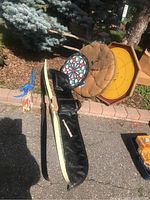 Photo showing two longbows with black zippered carry cases, about 62-64 inches long, lying on pavement with blue arrows with colorful fletching beside them. Target pad and dartboard visible in background leaning against tree.