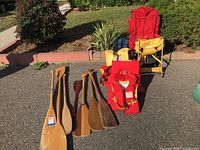 Full view of 7 wooden paddles, 6 life jackets, 3 backpacks including yellow infant backpack and red backpack, with one wooden toddler carrier in the background.
