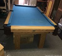Full side view of pool table showing light oak wood frame, blue felt surface, and leather drop pockets with fringe.