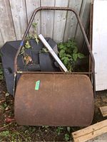 Front view of the rusty metal lawn roller and handle leaning against a shed with vines growing nearby.