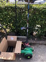 Overall view of the lot items including green Weed Eater leaf blower, Troy-Bilt gas trimmer, broom, and cardboard box with parts.