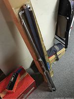 Tripod folded leaning against box and wall, possibly Velbon tripod or table top tripod in case, showing tripod legs and part of carrying case.