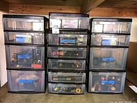 Three black plastic framed storage drawer units with multiple clear drawers containing various small hardware and gardening tools labeled with tape.