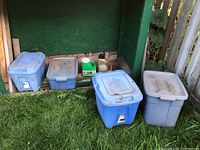 Four plastic storage bins containing yard chemicals outdoors on grass against green fence