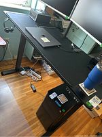 Full view of black metal adjustable computer desk with laptop and monitor on top, cables and power strip visible underneath.