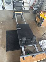 Aero Pilates reformer machine angled from the front, showing padded carriage with two cylindrical leg stops and metal frame with wooden end panel on the floor.