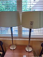 Two chrome floor lamps with white drum shades standing on a wooden surface near a window with blinds.