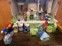 Photo of cleaning supplies on floor including multiple spray bottles, boxes of wipes, bleach, and sponges under a sink.