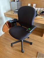 Front view of the black fabric office chair showing the cushioned seat, backrest, adjustable armrests, and caster wheels on hardwood floor near desk and filing drawers.