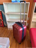 Red hard shell Delsey Paris suitcase in front of wooden shelving, showing front and side views with telescoping handle extended