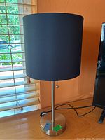 Full view of one chrome desk lamp with a black cylindrical shade placed on a wooden surface near a window.