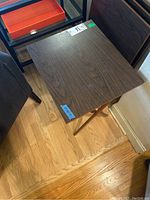 Photo showing one wooden folding dinner table with scratches on surface, dark wood grain finish, standing on wooden floor near other furniture.