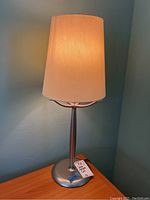 Full view of one silver tone table lamp lit, showing cylindrical fabric shade and round base on wooden surface