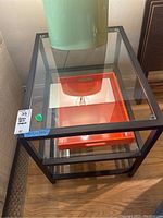 Front view of black metal framed glass topped square end table with green lamp on top and orange serving tray inside.