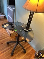 Glass office table with tinted glass top and black metal frame, on caster wheels, shown next to a wall and hardwood floor.