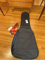 Black vinyl guitar case laid flat on wood floor next to floral patterned guitar strap with tags.
